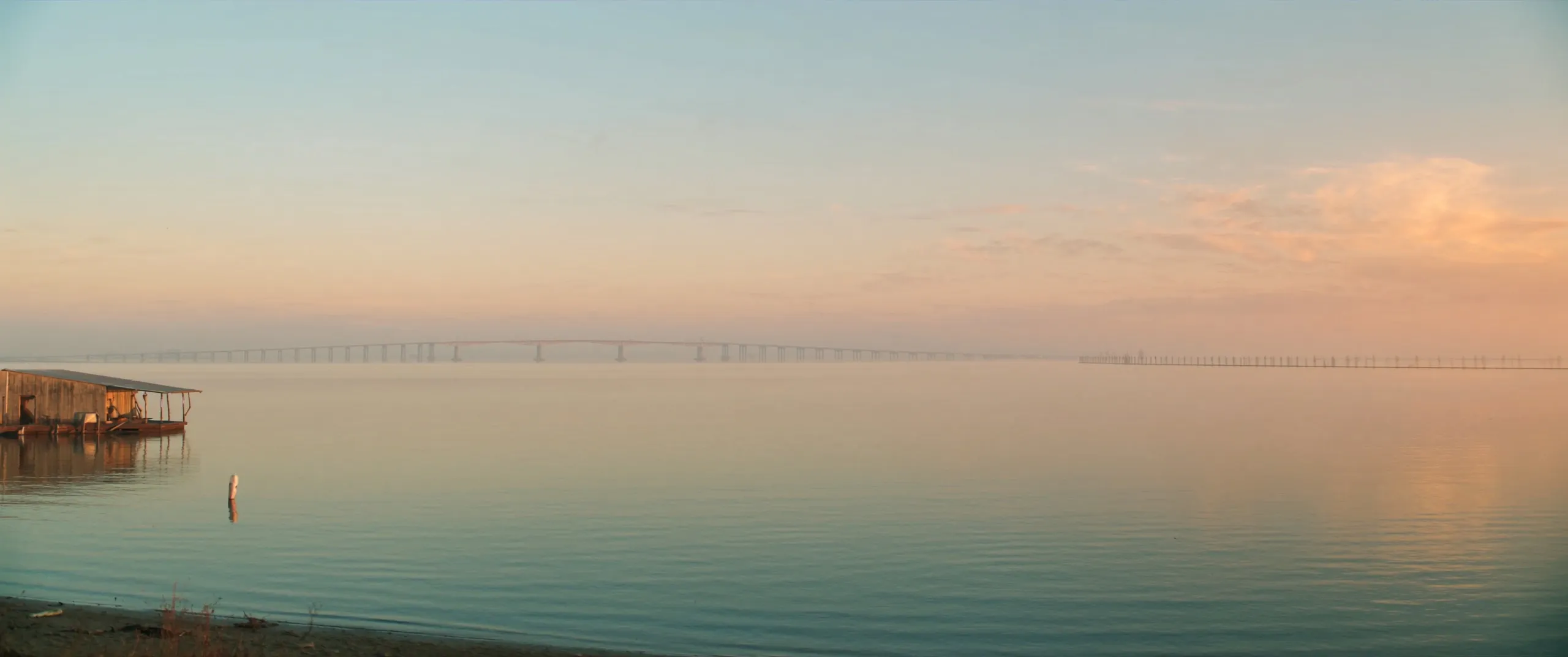 Lake Pontchartrain at dawn with the causeway faint on the horizon, seen from a quiet wooden shoreline